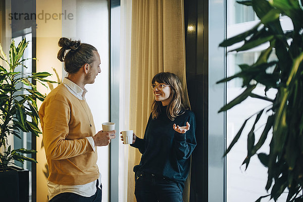 Smiling businesswoman and businessman talking while drinking by window at office