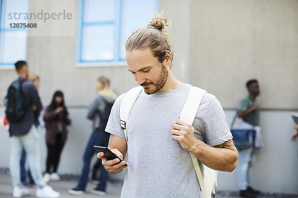 Male university student using mobile phone at campus with friends standing in background