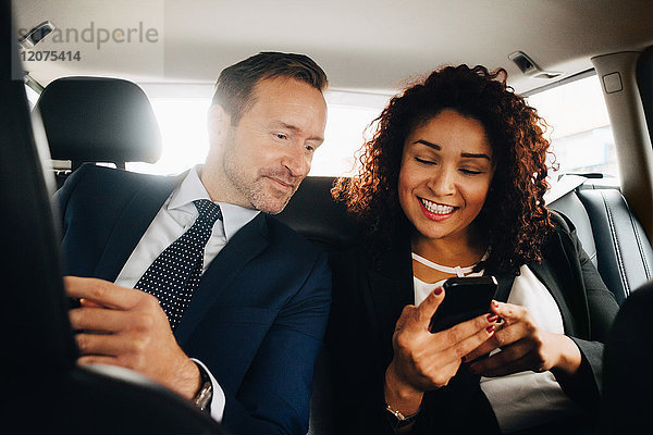 Smiling businessman colleagues sharing smart phone while sitting in taxi