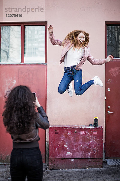 Rear view of teenage girl photographing female friend jumping against building