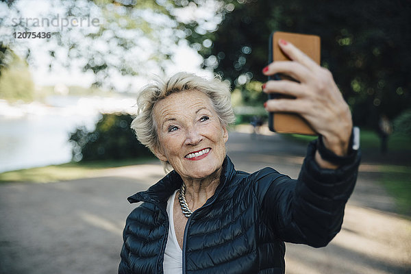 Lächelnde Seniorenfrau nimmt Selfie mit Smartphone in den Park