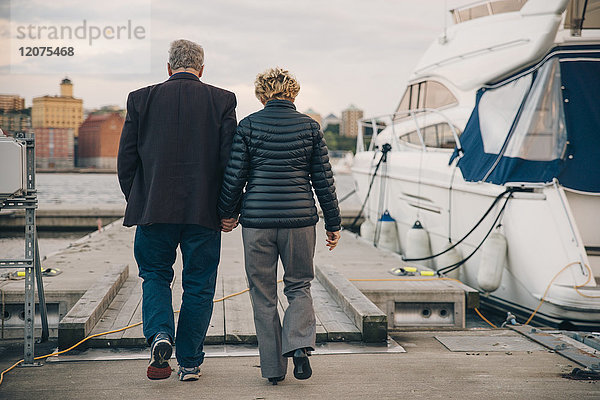 Durchgehende Rückansicht des älteren Paares  das beim Gehen auf dem Pier am Hafen Händchen hält