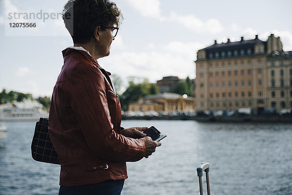 Seitenansicht einer älteren Frau  die ein Mobiltelefon hält  während sie in der Stadt am Meer steht.