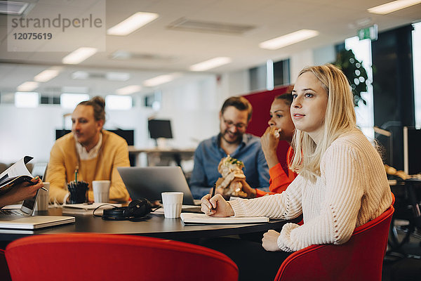 Nachdenkliche junge Geschäftsfrau schaut weg  während sie mit Kollegen am Konferenztisch im Büro sitzt.