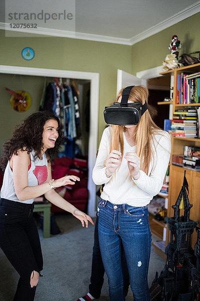 Fröhliches Teenagermädchen  das einen Freund mit einem Virtual-Reality-Headset zu Hause ansieht.