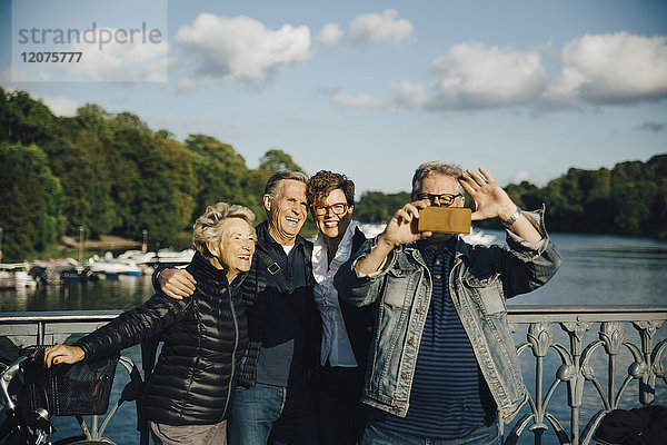 Lächelnde männliche und weibliche Freunde nehmen Selfie mit Smartphone durch Geländer von Brücke