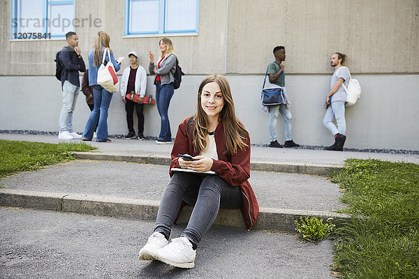 Porträt einer lächelnden Frau  die auf dem Uni-Campus mit Studenten im Hintergrund sitzt.
