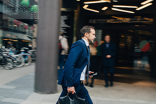 Seitenansicht des reifen Geschäftsmannes mit Tasche und Smartphone auf dem Bürgersteig in der Stadt