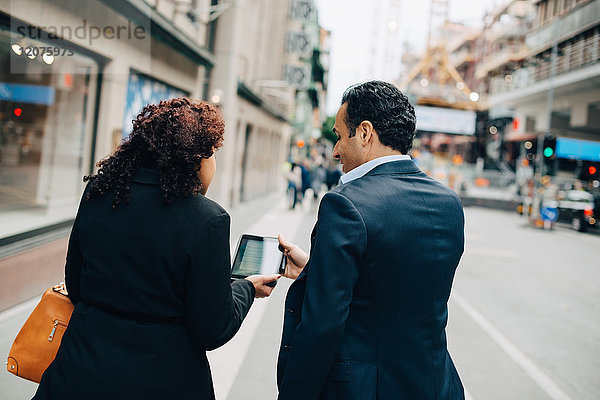 Rückansicht der Geschäftsfrau  die dem Geschäftsmann beim Gehen auf dem Bürgersteig in der Stadt ein digitales Tablett zeigt.