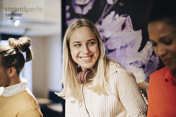 Lächelnde blonde Geschäftsfrau mit Kopfhörer von Geschäftsmann  die eine Kollegin im Büro anschaut.