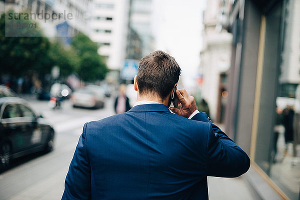 Rückansicht eines reifen Geschäftsmannes  der auf dem Handy spricht  während er in der Stadt auf dem Bürgersteig läuft.