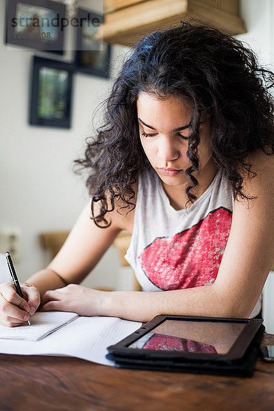 Teenager-Mädchen studiert am Tisch beim Schreiben in Buch durch digitale Tablette