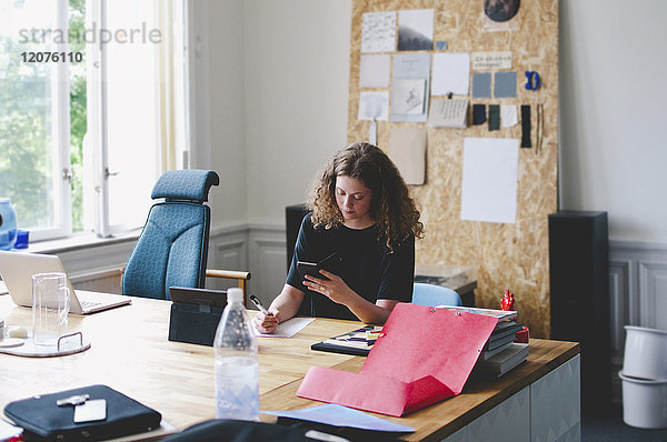 Junge Geschäftsfrau mit Handy beim Schreiben auf Papier am Schreibtisch im Kreativbüro