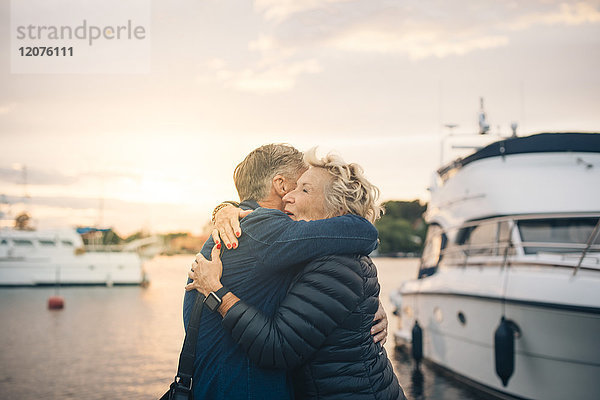 Seitenansicht eines älteren Mannes und einer älteren Frau  die sich bei Sonnenuntergang am Hafen umarmen.