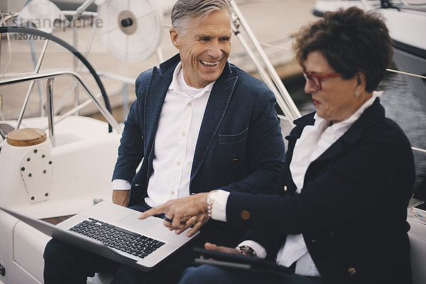 Lächelnder älterer Mann  der auf eine Frau schaut  die auf den Laptop zeigt  während sie auf einer Yacht unterwegs ist.