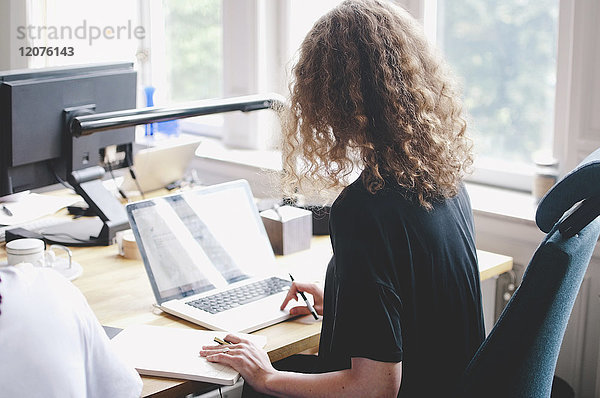 Rückansicht der jungen Geschäftsfrau mit Laptop am Tisch im Kreativbüro