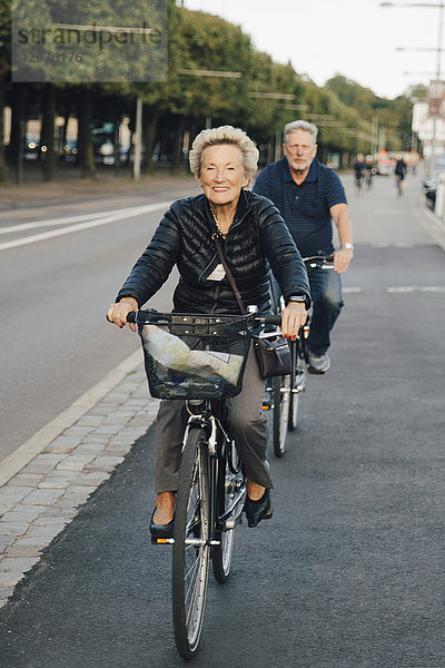 Lächelnde Seniorenfrau mit Mann beim Fahrradfahren auf der Stadtstraße