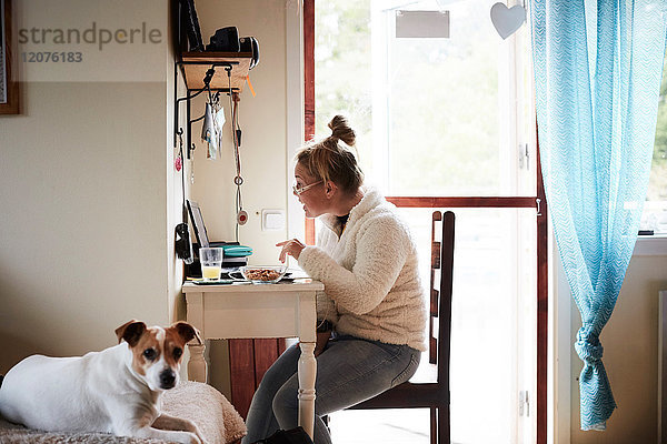 Behinderte junge Frau mit Laptop am Schreibtisch mit Hund im Vordergrund zu Hause