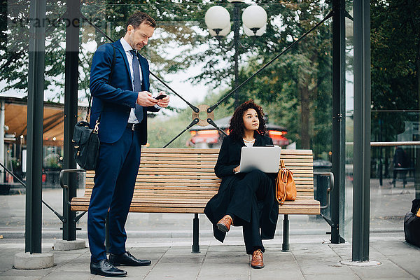 Volle Länge der Geschäftsleute  die Smartphones benutzen  während sie an der Bushaltestelle warten.