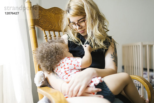 Mutter sitzt auf einem Stuhl und stillt ihre Tochter (12-17 Monate)