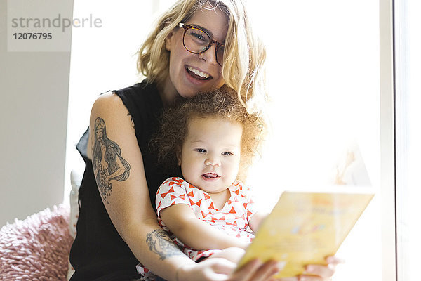 Mutter sitzt mit Tochter (12-17 Monate) auf der Fensterbank und liest ein Buch