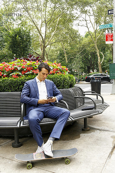 Kaukasischer Geschäftsmann auf einer Parkbank  der eine SMS auf seinem Mobiltelefon schreibt