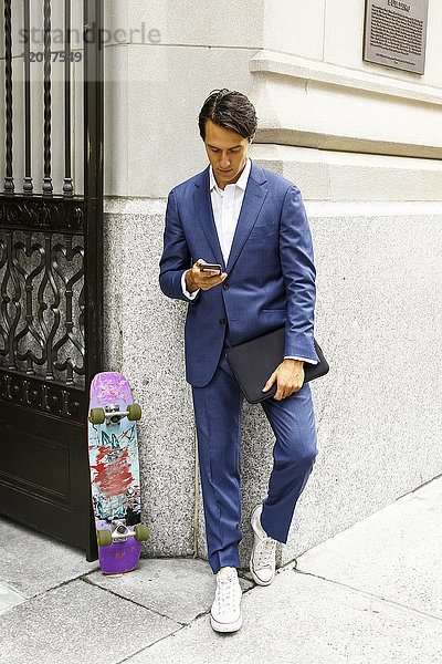 Kaukasischer Geschäftsmann mit Skateboard  der eine SMS auf seinem Mobiltelefon schreibt