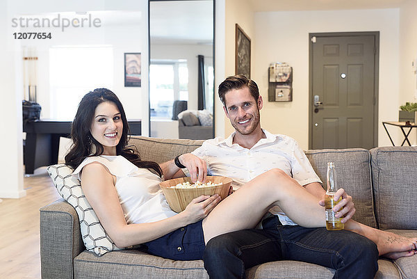 Lächelndes kaukasisches Paar entspannt sich auf dem Sofa mit Bier und Popcorn