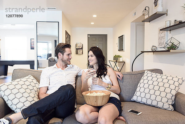 Lächelndes kaukasisches Paar entspannt sich auf dem Sofa mit Wein und Popcorn