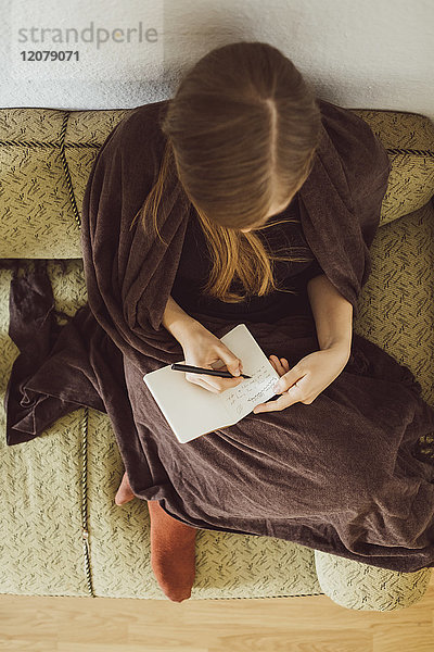 Frau sitzt auf der Couch im Wohnzimmer und schreibt in Notizbuch  Draufsicht