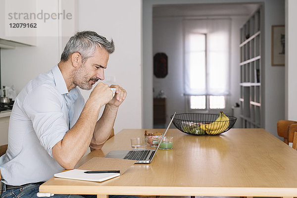 Lächelnder Mann trinkt Kaffee und benutzt Laptop im Home-Office