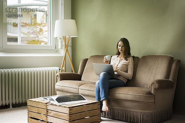 Junge Frau sitzend auf der Couch mit Tasse Kaffee mit Laptop