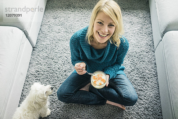 Porträt einer glücklichen Frau mit einem Hund  der Fruchtjoghurt im Wohnzimmer isst.