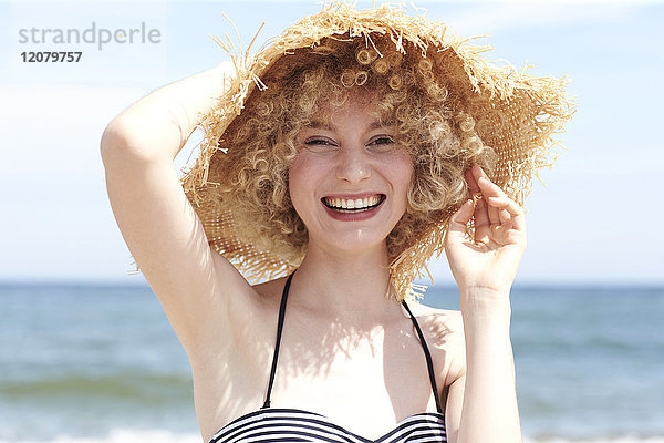 Porträt einer lachenden jungen Frau mit Strohhut am Strand
