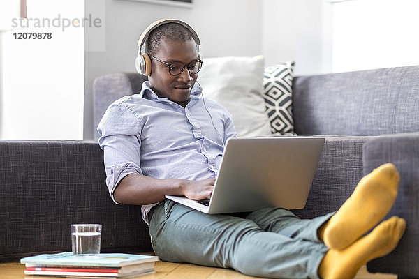 Junger Mann sitzt auf dem Boden im Wohnzimmer mit Laptop und Kopfhörer