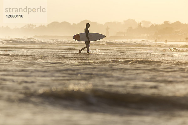 Indonesien  Bali  Surfer  der sein Surfbrett bei Sonnenuntergang ins Meer trägt.