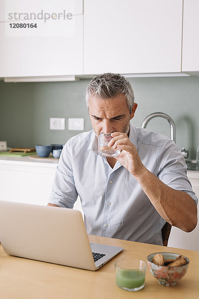 Mann trinkt Wasser aus Glas und nutzt Laptop im Home-Office