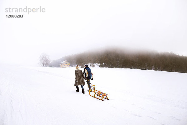 Rückansicht des Seniorenpaares mit Schlittenwanderung nebeneinander in verschneiter Landschaft