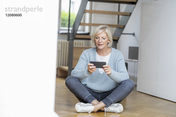 Porträt einer älteren Frau  die zu Hause auf dem Boden sitzt und ein Smartphone benutzt.