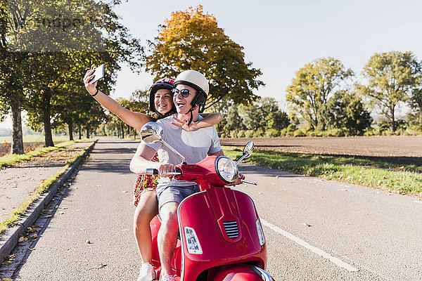 Glückliches junges Paar mit einem Selfie auf dem Motorroller auf der Landstraße