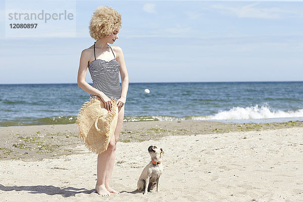 Junge Frau im Badeanzug mit Strohhut und Hund am Strand