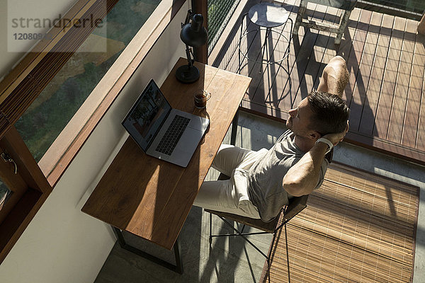 Mann lehnt sich zurück und macht eine Pause von der Arbeit am Laptop auf dem Schreibtisch im Haus.