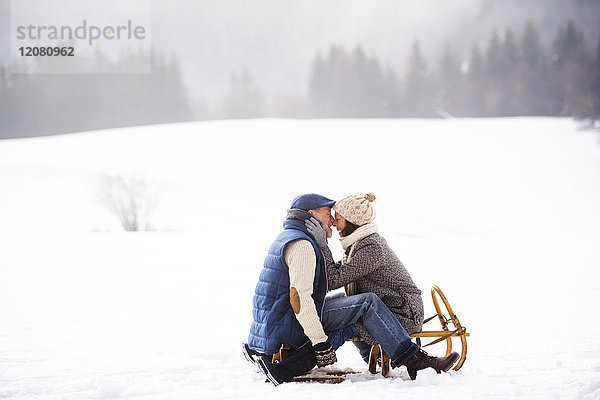 Glückliches Seniorenpaar sitzt von Angesicht zu Angesicht auf einem Schlitten in der Winterlandschaft.
