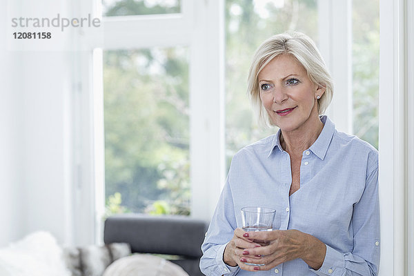 Porträt einer lächelnden Seniorin mit einem Glas Wasser im Wohnzimmer