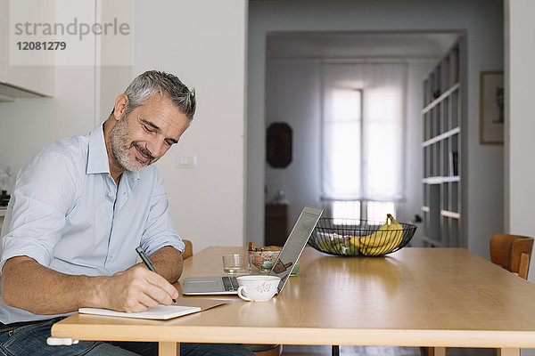 Lächelnder Mann beim Schreiben im Notebook und Laptop im Home-Office