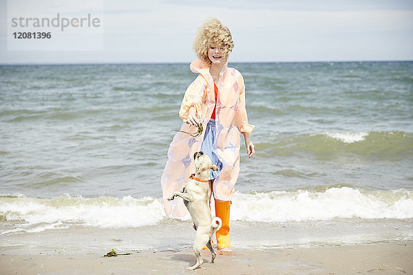 Lächelnde junge Frau spielt mit ihrem Hund am Strand