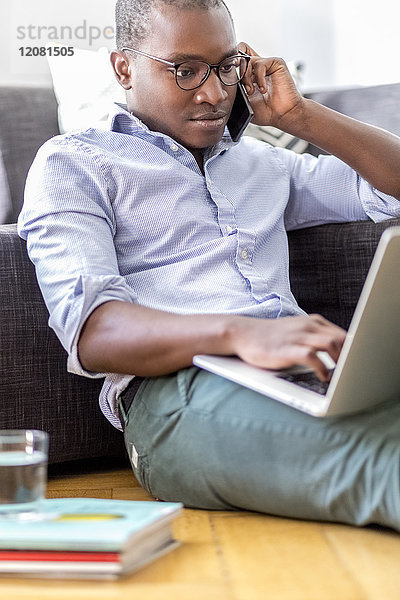 Junger Mann am Telefon sitzt auf dem Boden im Wohnzimmer mit Laptop