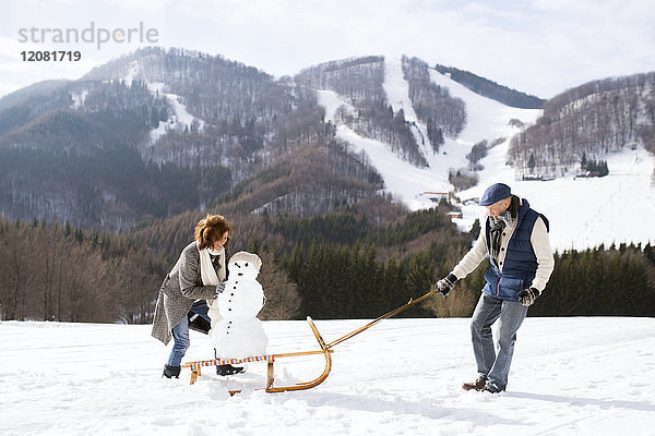 Seniorenpaar mit Schneemann auf Schlitten in Winterlandschaft