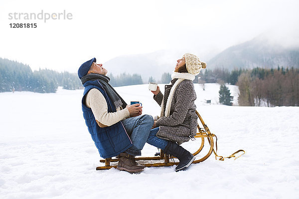 Seniorenpaar auf Schlitten sitzend mit heißen Getränken in verschneiter Winterlandschaft nach oben schauend