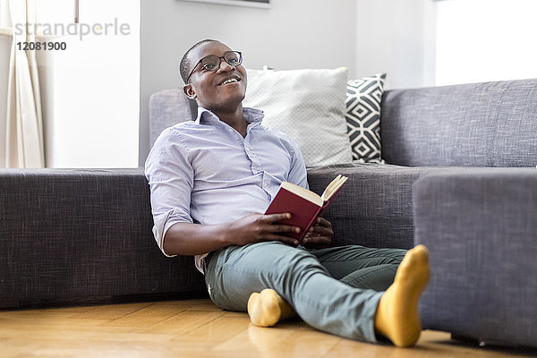 Glücklicher junger Mann sitzt auf dem Boden im Wohnzimmer und liest das Buch.